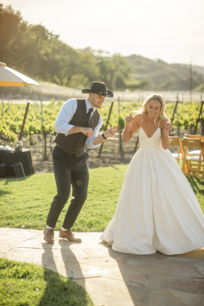 A newly wed couple dances up the walk to their wedding reception. The man is wearing his suit and cowboy hat. The woman is wearing a lovely, white wedding gown.