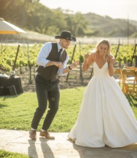 A newly wed couple dances up the walk to their wedding reception. The man is wearing his suit and cowboy hat. The woman is wearing a lovely, white wedding gown.