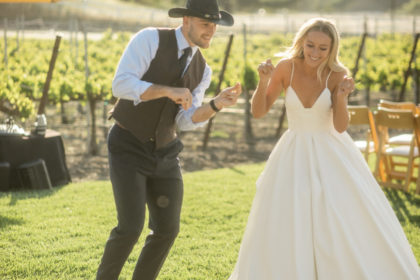 A newly wed couple dances up the walk to their wedding reception. The man is wearing his suit and cowboy hat. The woman is wearing a lovely, white wedding gown.